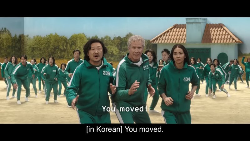 Will Ferrell is shown in a scene from Squid Game along with other individuals, all wearing the same green tracksuit. Open subtitles read, "You moved!" Closed captions at the very bottom of our screen read, "[In Korean] You moved."