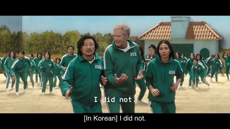 Will Ferrell is shown in a scene from Squid Game along with other individuals all wearing the same green tracksuit. Open subtitles read, "I did not." Closed captions at the very bottom of our screen read, "[In Korean] I did not."