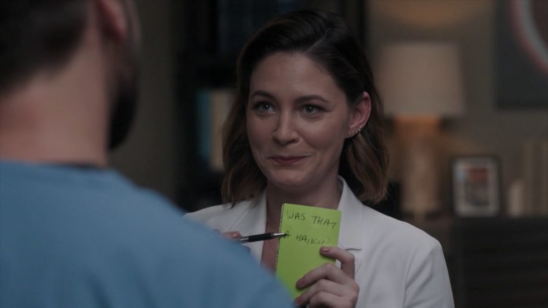 Elizabeth, wearing a hospital coat and smiling as she stands by Max, is holding a green piece of paper that reads "Was that a haiku?" She also has a pen pointing to the question on the paper.