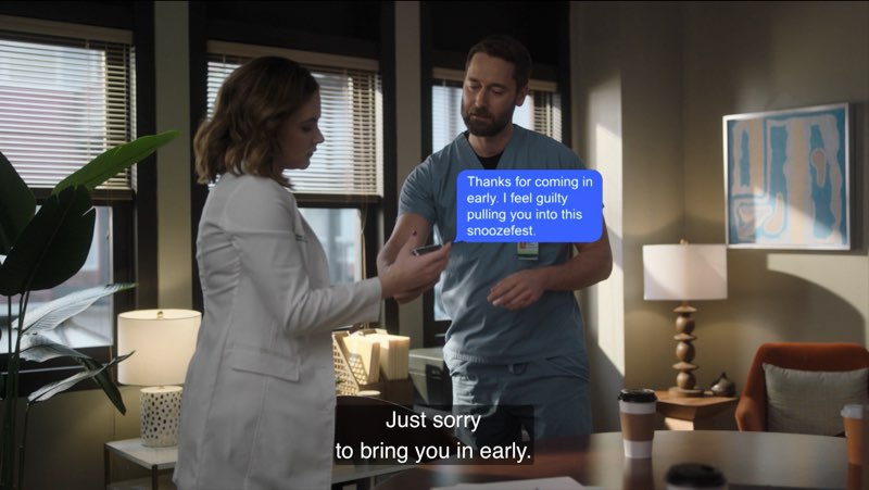 Max and Elizabeth are standing near each other in her hospital office. He holds a phone out for her to look at. A blue bubble appears near them that reads, "Thanks for coming in early. I feel guilty pulling you into the snoozefest." Closed captions at the bottom state, "Just sorry to bring you in early."