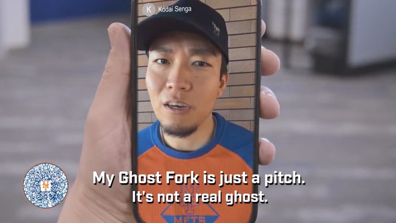 Kodai Senga is shown on a smartphone that someone is holding. He is wearing the Mets colors of blue and orange, and a baseball cap. He is speaking and subtitles appear at the bottom of our screen reading, "My Ghost Fork is just a pitch. It's not a real ghost."
