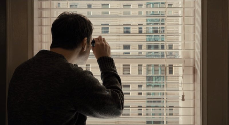 Theo uses a pair of binoculars to peer through his apartment window into another apartment window.