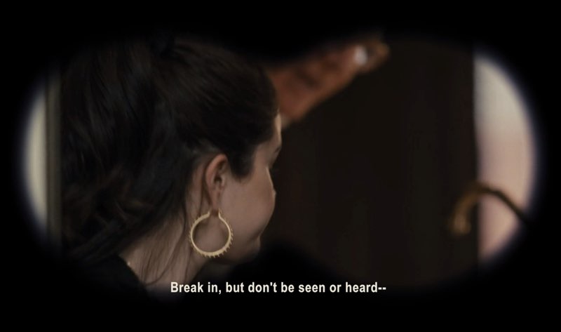 Mabel and Charles are seen through Theo’s binoculars. Charles is talking, open captions at the bottom reading, “Break in, but don’t be seen or heard—”