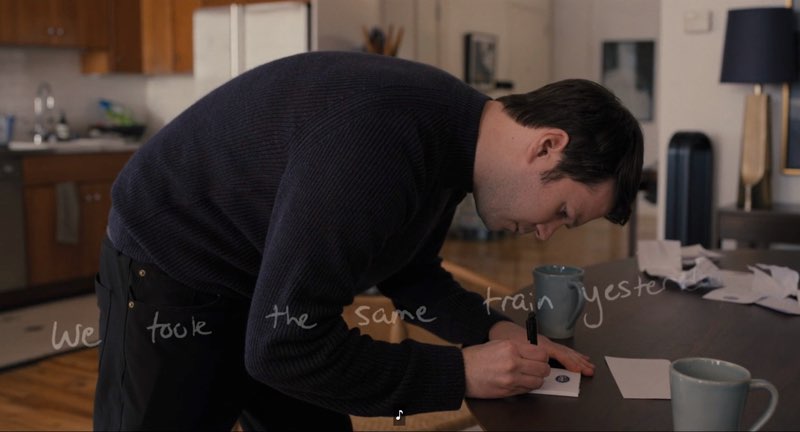 Theo is in his apartment, hunched over a table writing on a notepad. The handwritten words are shown across the screen saying, "We took the same train yesterday."