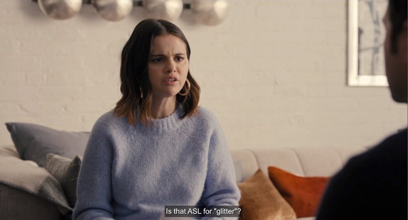 Mabel is seated in front of Theo in his apartment as she speaks. Closed captions appear at the bottom: "Is that ASL for "glitter?"