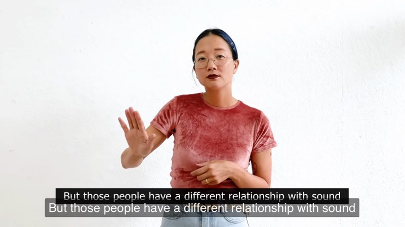 Christine Sun Kim, standing against a white background, signs to the camera. The same line is captioned twice at the bottom of the screen and reads, "But those people have a different relationship with sound."