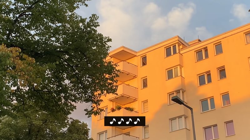 An apartment building is shown against a blue sky next to a tree. One line of captions appears at the bottom of the screen. This caption line shows four musical notes in a row.