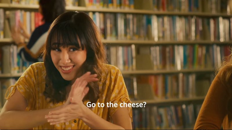A woman student is in a library signing, “cheese?” with a questioning look on her face. Subtitles at the bottom read, "Go to the cheese?"