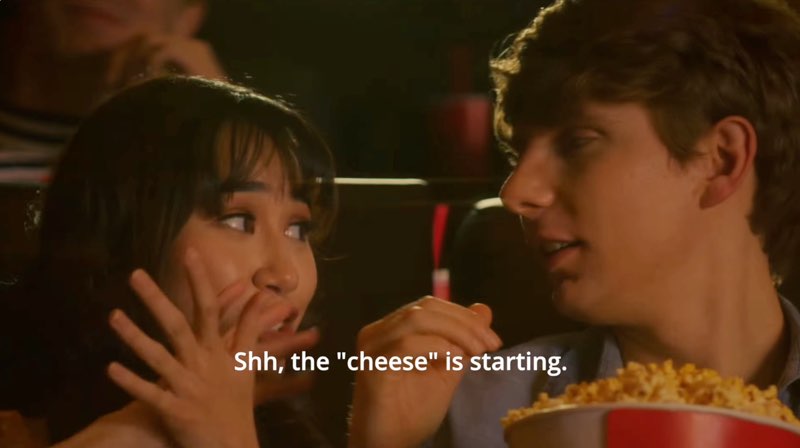 The young man and woman are seated in a movie theater with popcorn as they look at each other. The woman is signing the word, “cheese.” Subtitles at the bottom read, "Shh, the ‘cheese’ is starting."