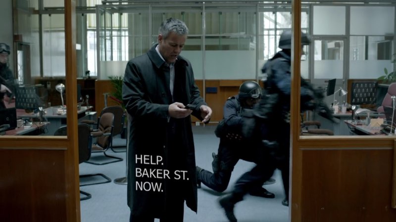 Lestrade, standing in the middle of an office, stares concernedly at his phone. Visual text appears in the space below his phone: "HELP. BAKER ST. NOW."