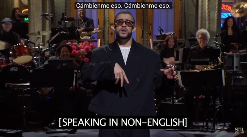 Bad Bunny is standing on stage in front of the “Saturday Night Live” house band. He is gesturing to open subtitles at the bottom that read, "[Speaking in non-English]." Closed captions appear at the top of the screen in Spanish: “Cámbienme eso. Cámbienme eso.”