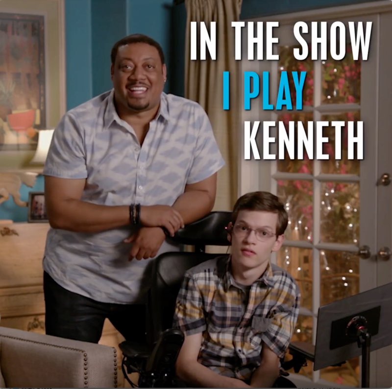 Cedric stands next to Micah, a white young male with short hair and glasses, says with open captions next to him, "In the show I play." The first line is in white and the second line is in blue.