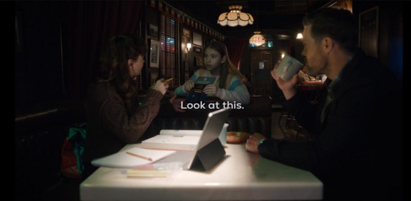 Ollie, Birdie, and her father are seated around a table at the bar. Ollie holds a document, showing it to Birdie. Subtitles appear underneath Ollie's hands that say, "Look at this."