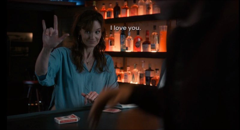 Birdie, smiling, is behind the bar counter, signing, "I love you" with one hand. Subtitles appear next to her near the center of the screen that read, "I love you."